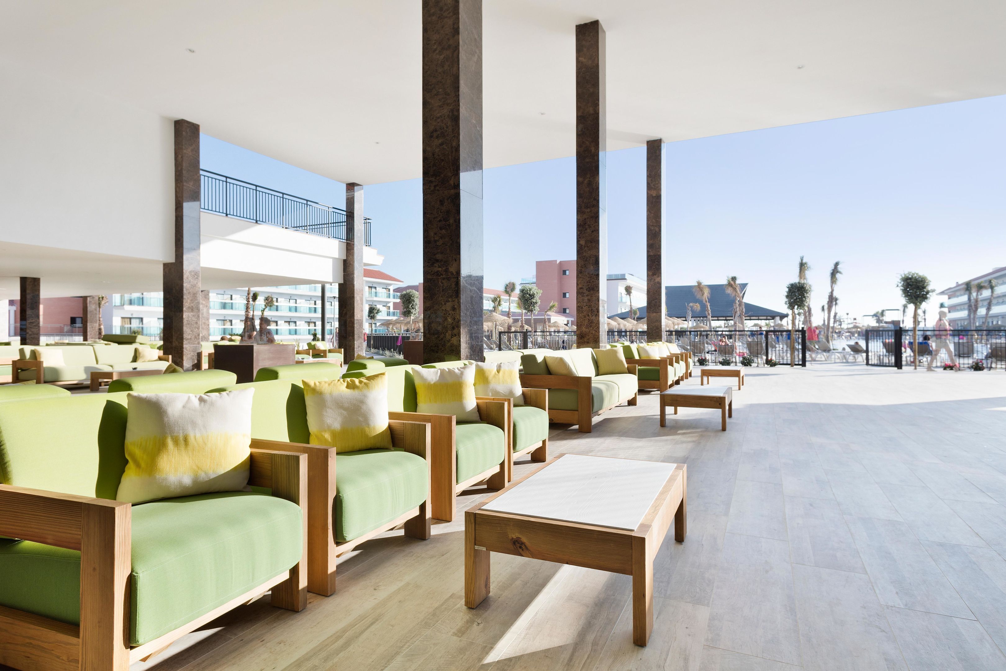Un área de descanso moderna con sofás verdes y mesas de madera, grandes pilares, y vista a la playa y edificios al fondo.