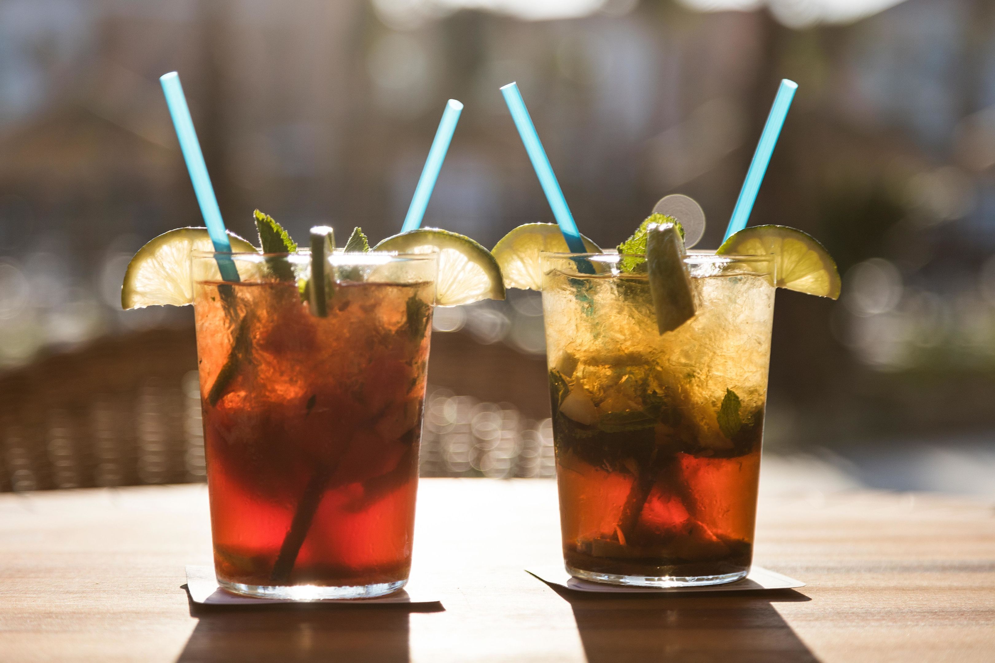 Two glasses of iced drinks with lime slices and mint leaves, each with a blue straw, on a wooden table.
