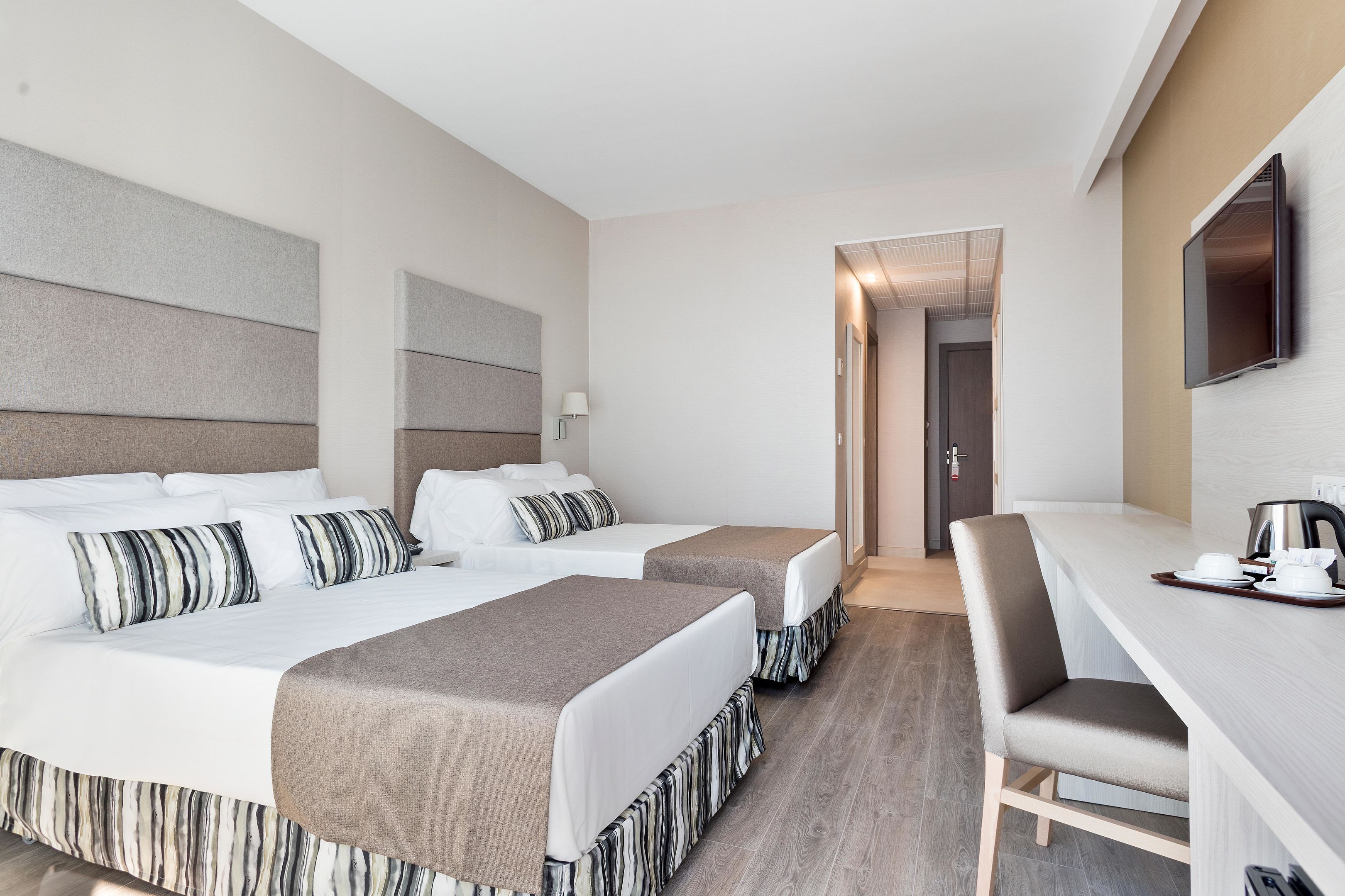 Habitación de hotel con dos camas, un escritorio, una silla, un televisor y una puerta que da a otra habitación.