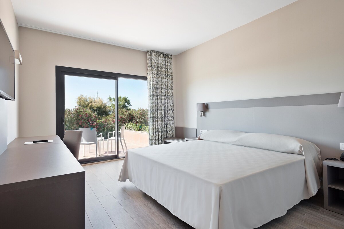 Ein modernes Hotelzimmer mit einem großen Bett, einem Schreibtisch und einem Balkon mit Blick auf Bäume und Blumen.