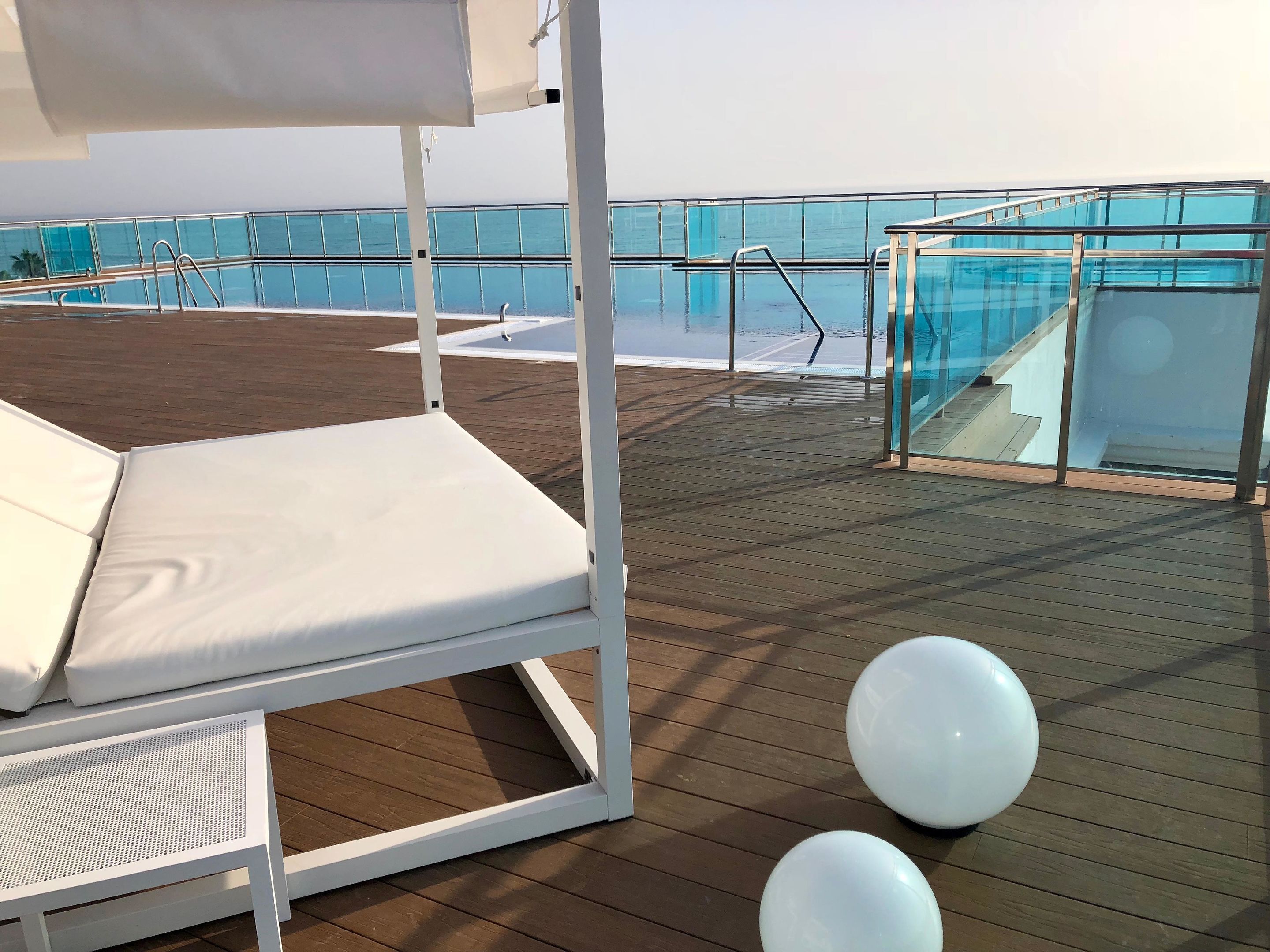 Terraza iluminada por el sol con una silla de descanso blanca, barandilla de cristal y zona de piscina.