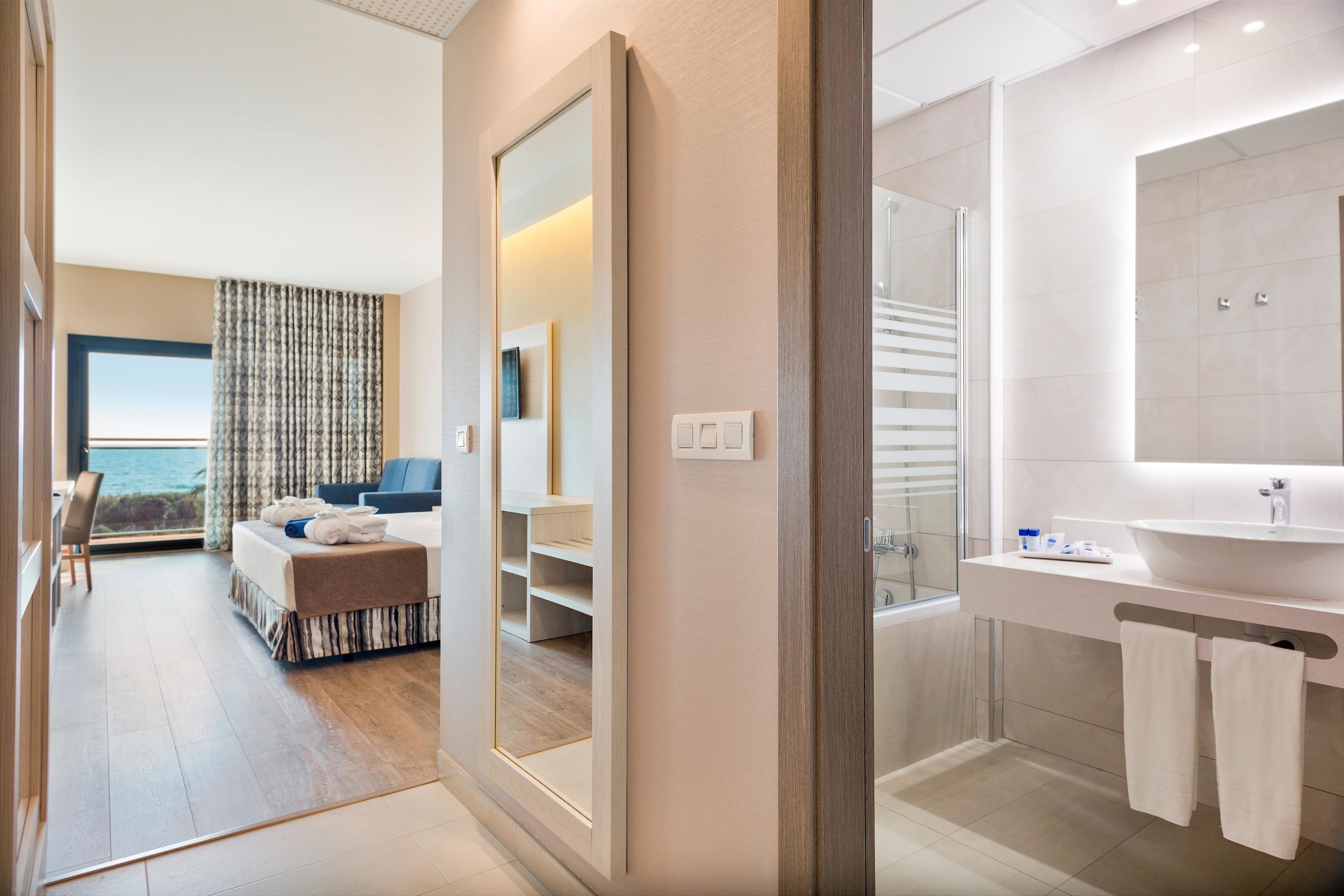 Habitació d'hotel moderna amb un llit gran, zona de seients, bany amb un mirall gran i un lavabo, i vista al mar per la finestra.