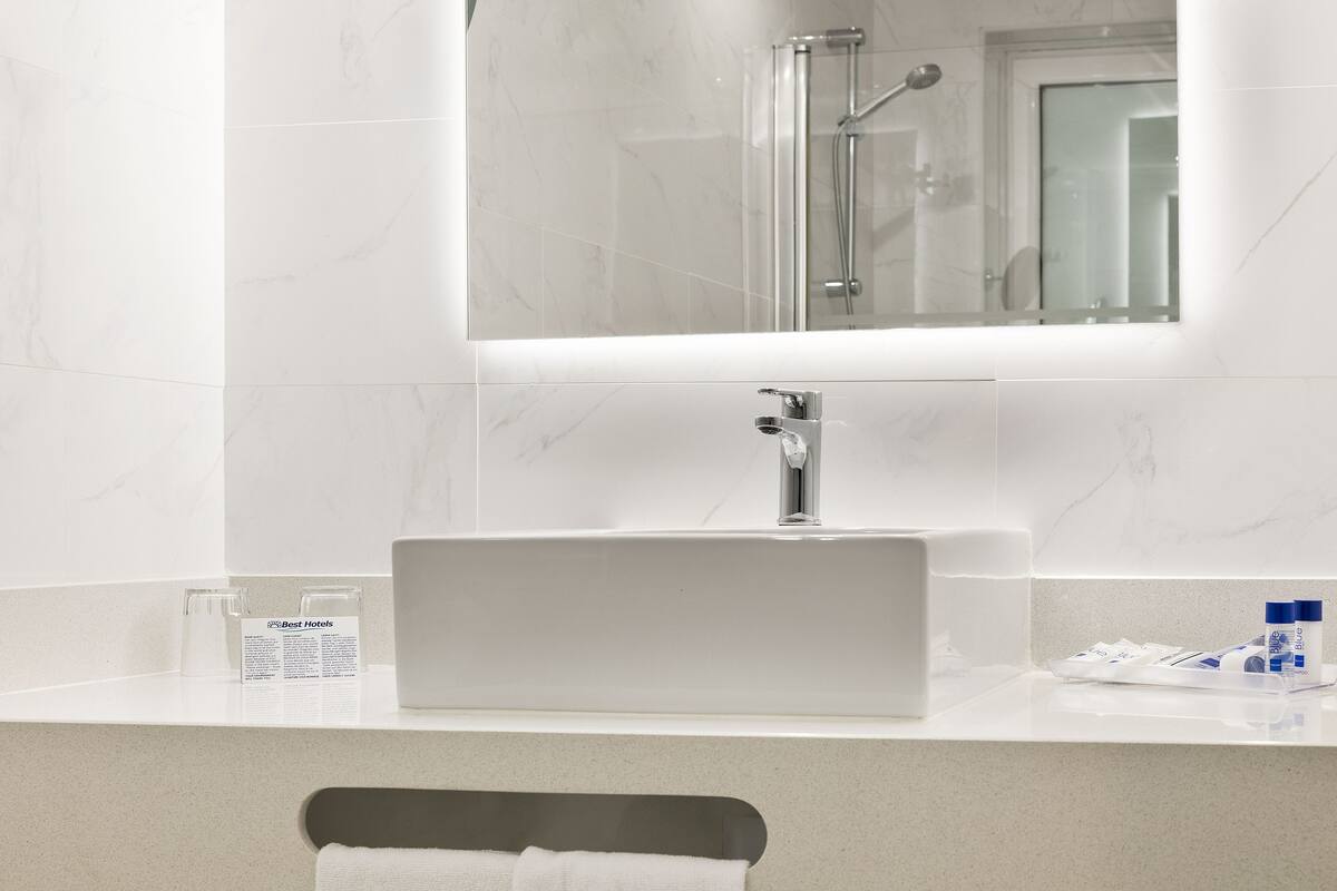 A bathroom with a white sink, a mirror, and a shower. There is a towel rack and a small table with toiletries.