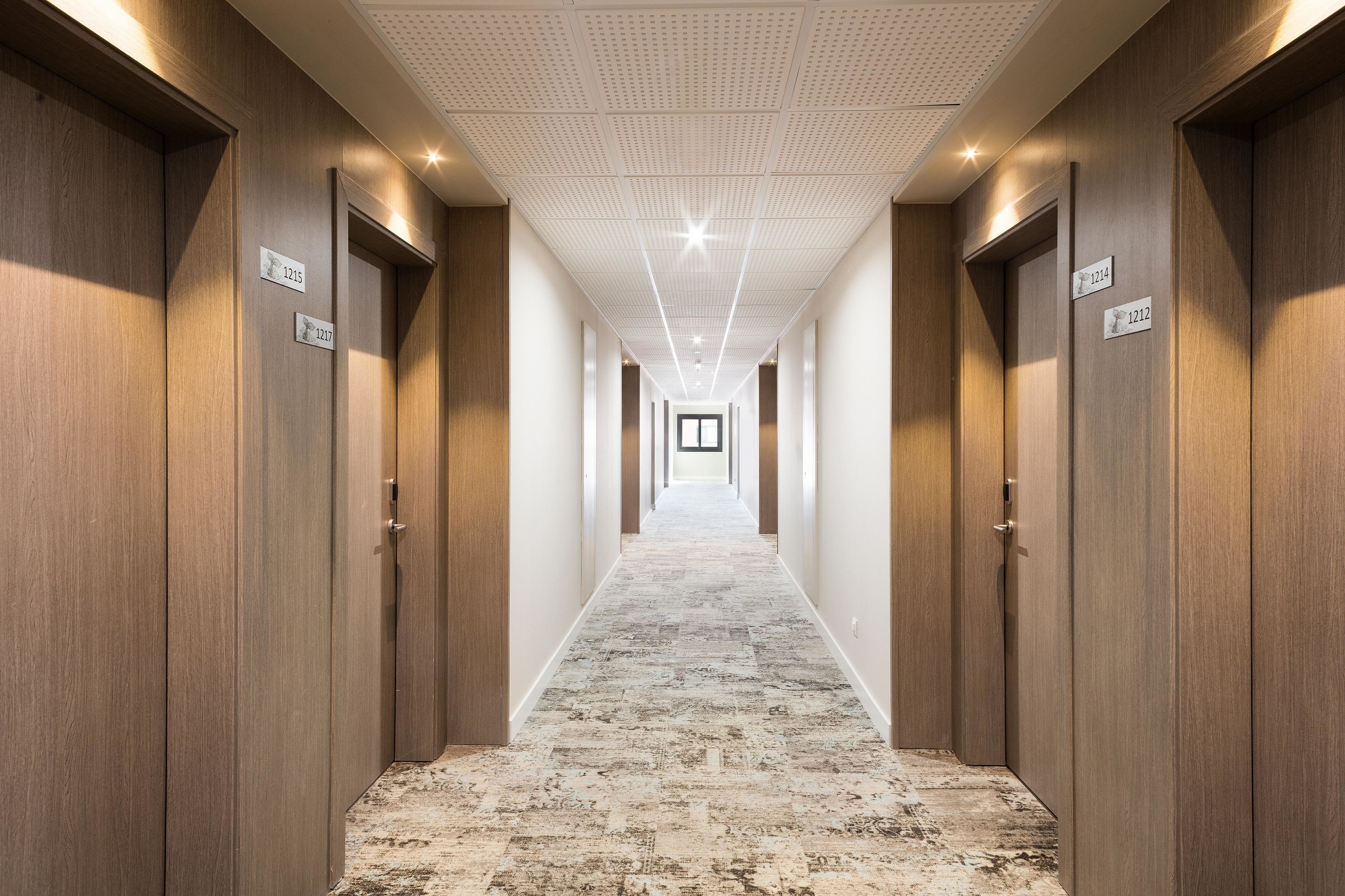 Pasillo de hotel con puertas de madera, letreros de habitaciones numeradas y una alfombra estampada.