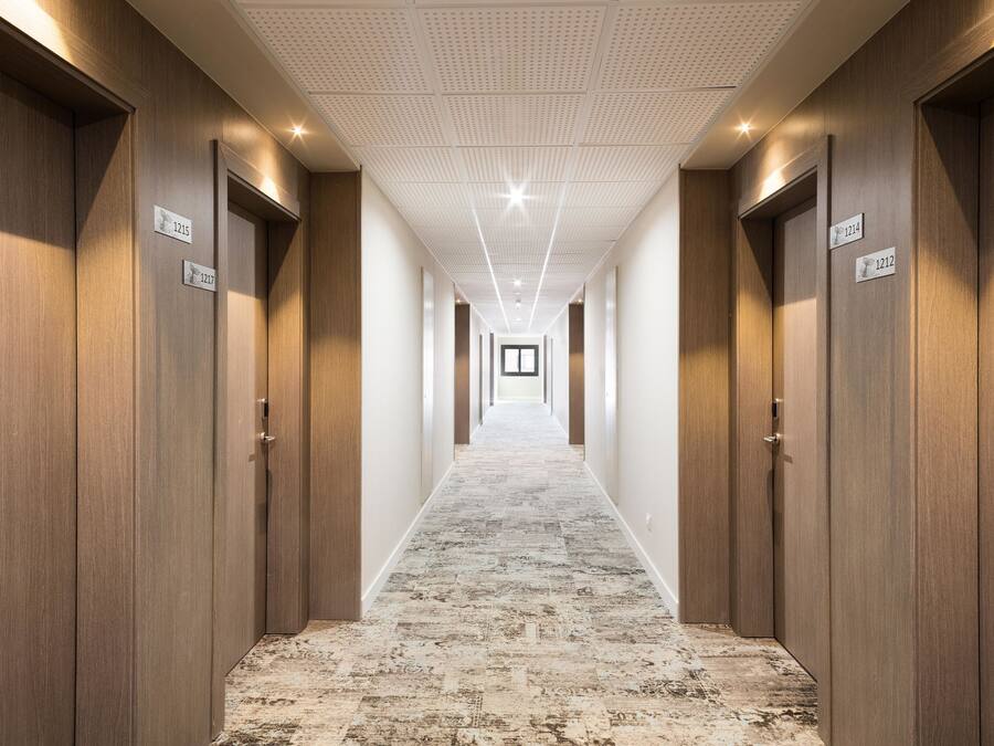 Pasillo de hotel con puertas de madera, letreros de habitaciones numeradas y una alfombra estampada.