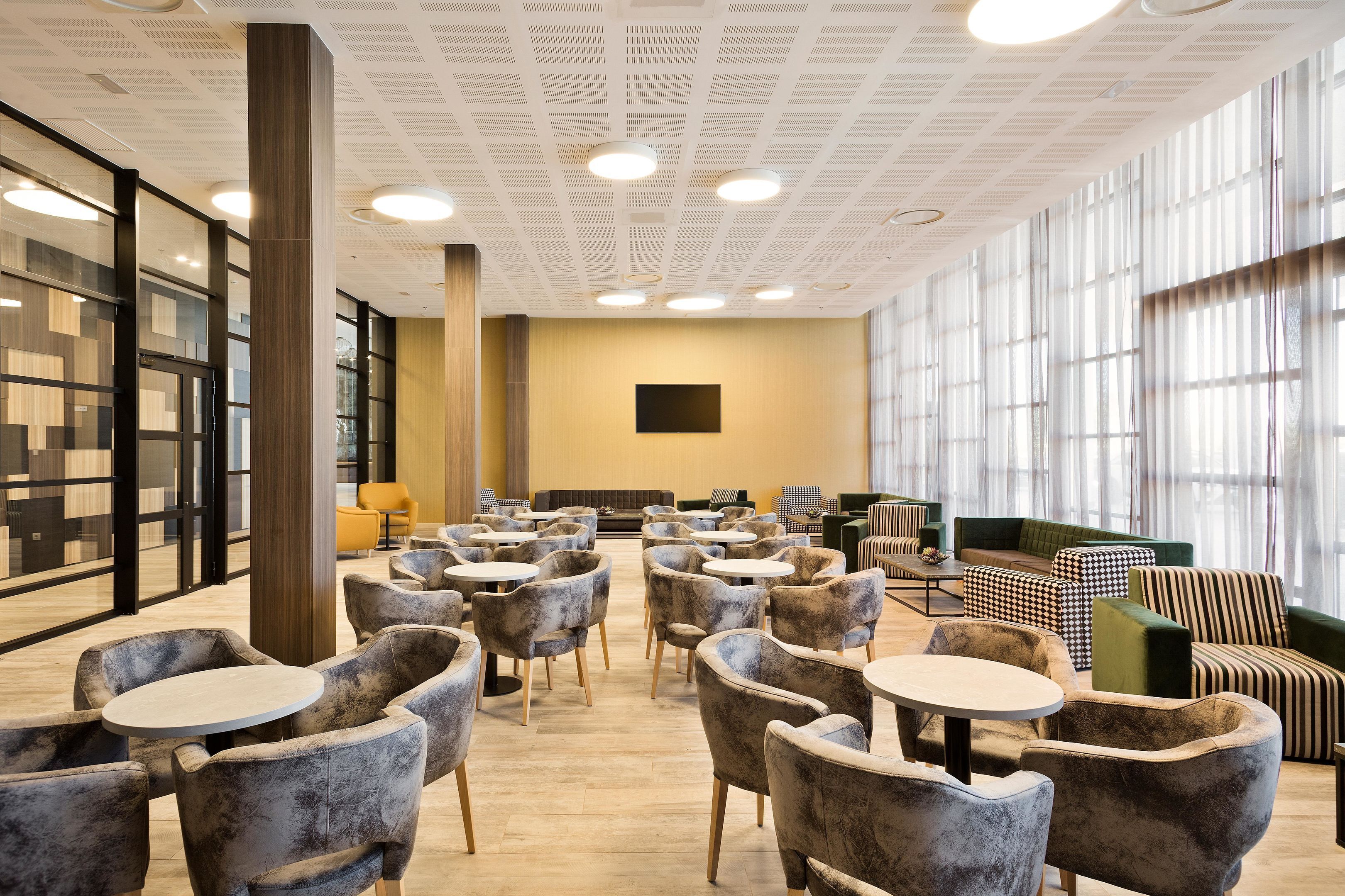A modern lounge area with round tables and chairs, a sofa, and a large window providing natural light.