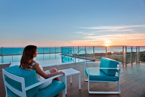 A woman sitting on a deck chair overlooking a pool and ocean at sunset.
