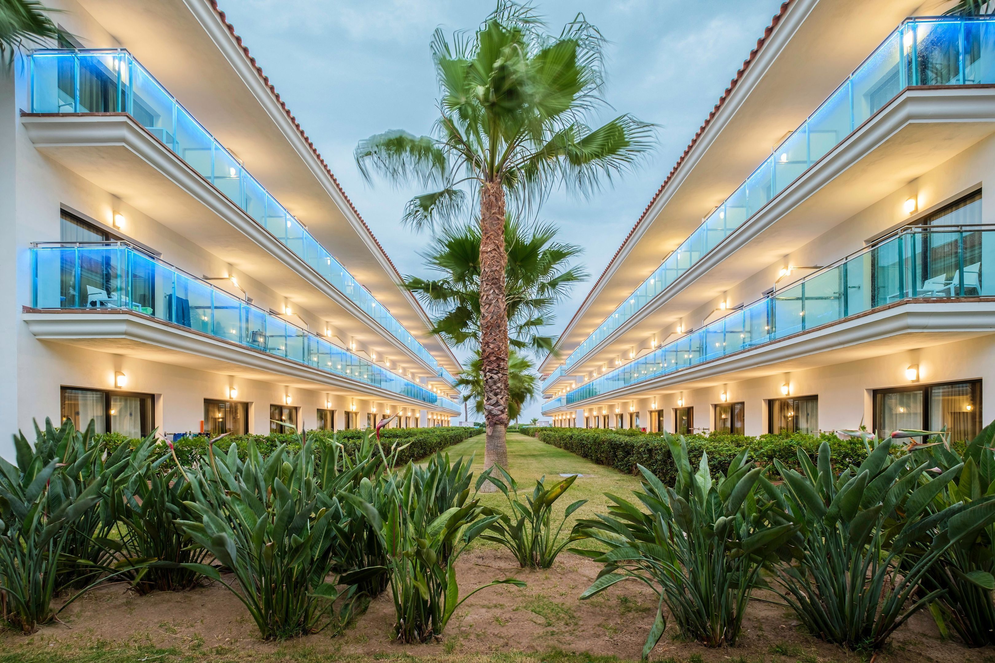 Un hotel moderno con un diseño simétrico, que presenta varios balcones con barandillas de vidrio e iluminación cálida.