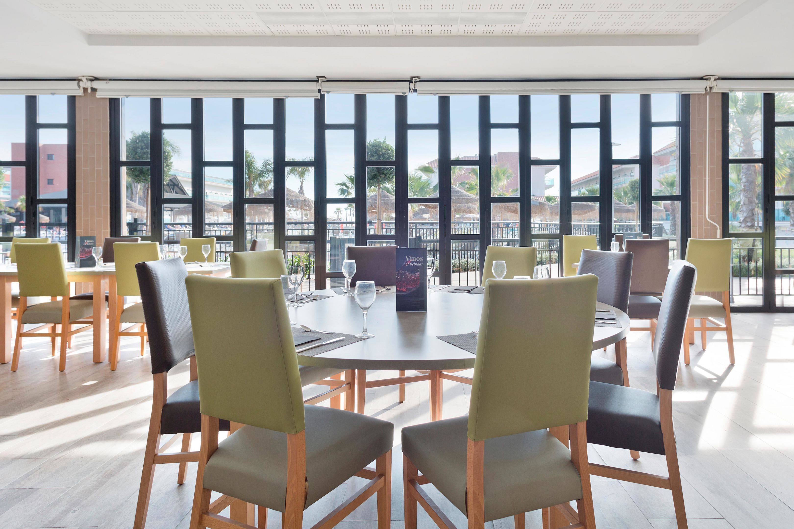 Un espace de restauration moderne avec des tables et des chaises rondes, de grandes fenêtres offrant une vue sur les immeubles et les palmiers.