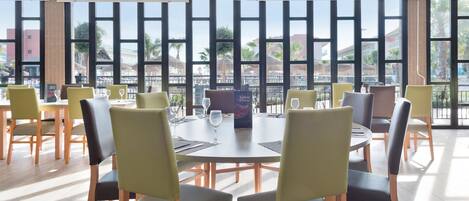 Un espace de restauration moderne avec des tables et des chaises rondes, de grandes fenêtres offrant une vue sur les immeubles et les palmiers.
