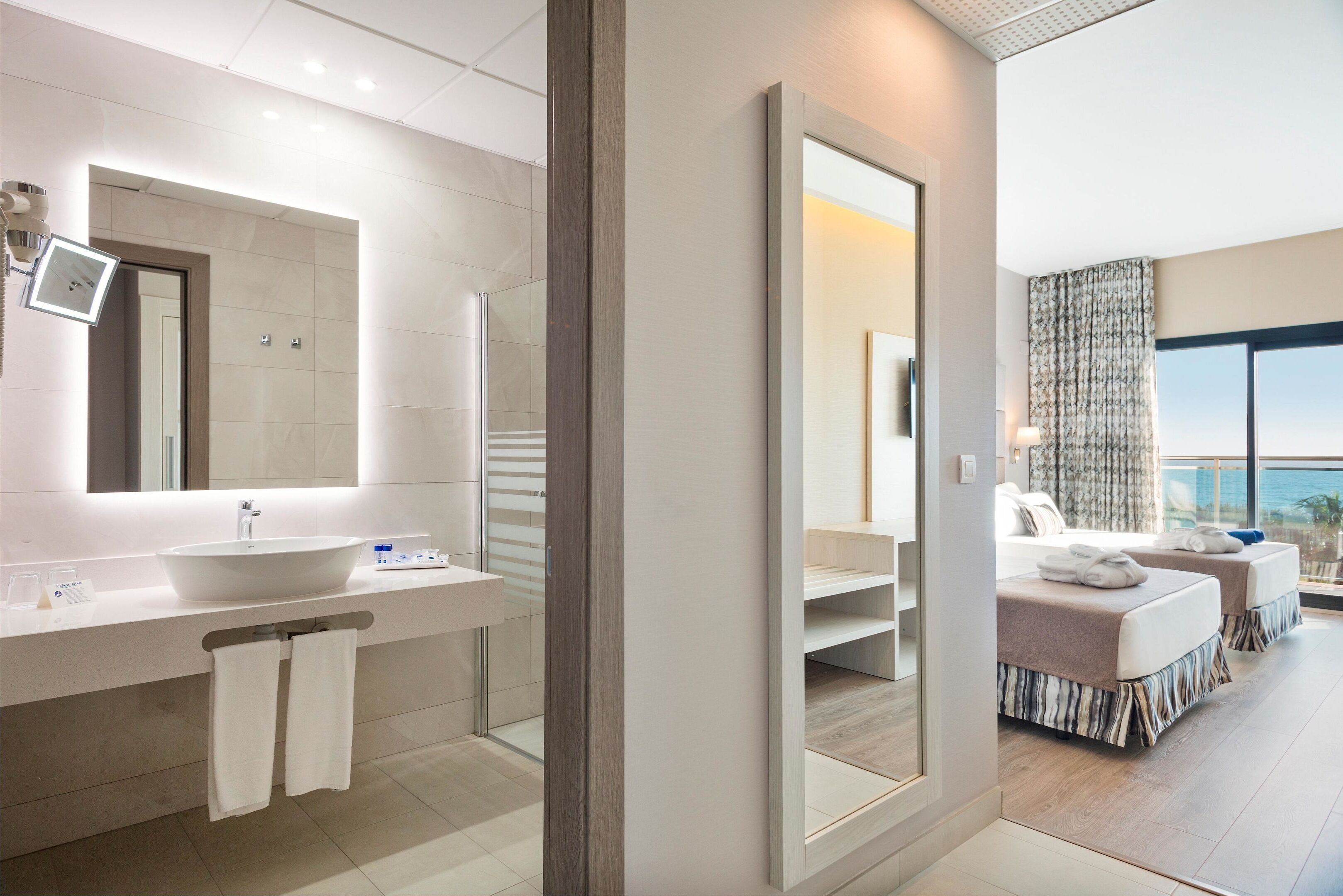 Un baño de hotel moderno con un lavamanos grande, un espejo y vistas al dormitorio con dos camas.