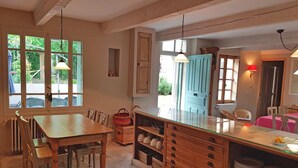 Une cuisine avec des meubles de rangement en bois, une table à manger et des chaises. On y trouve des fenêtres avec des rideaux, un luminaire suspendu et une porte avec un voyant rouge.