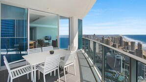 Un balcón moderno con vista a la ciudad y al océano, que incluye una zona de comedor con mobiliario blanco y una barandilla de vidrio.