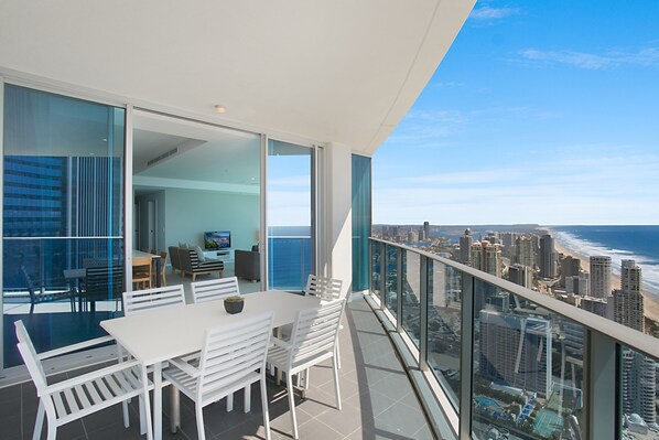 A modern balcony with a city and ocean view, featuring a dining area with white furniture and a glass partition.