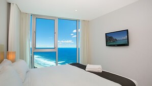 Habitación de hotel con una cama grande, un televisor en la pared y vistas al océano a través de un amplio ventanal.