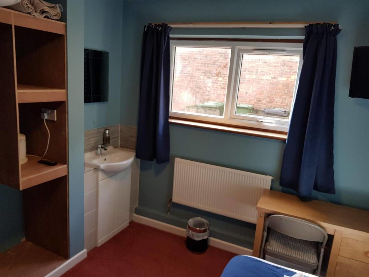 Una habitación pequeña y funcional con ventana, radiador, lavabo, silla y estante con espejo.