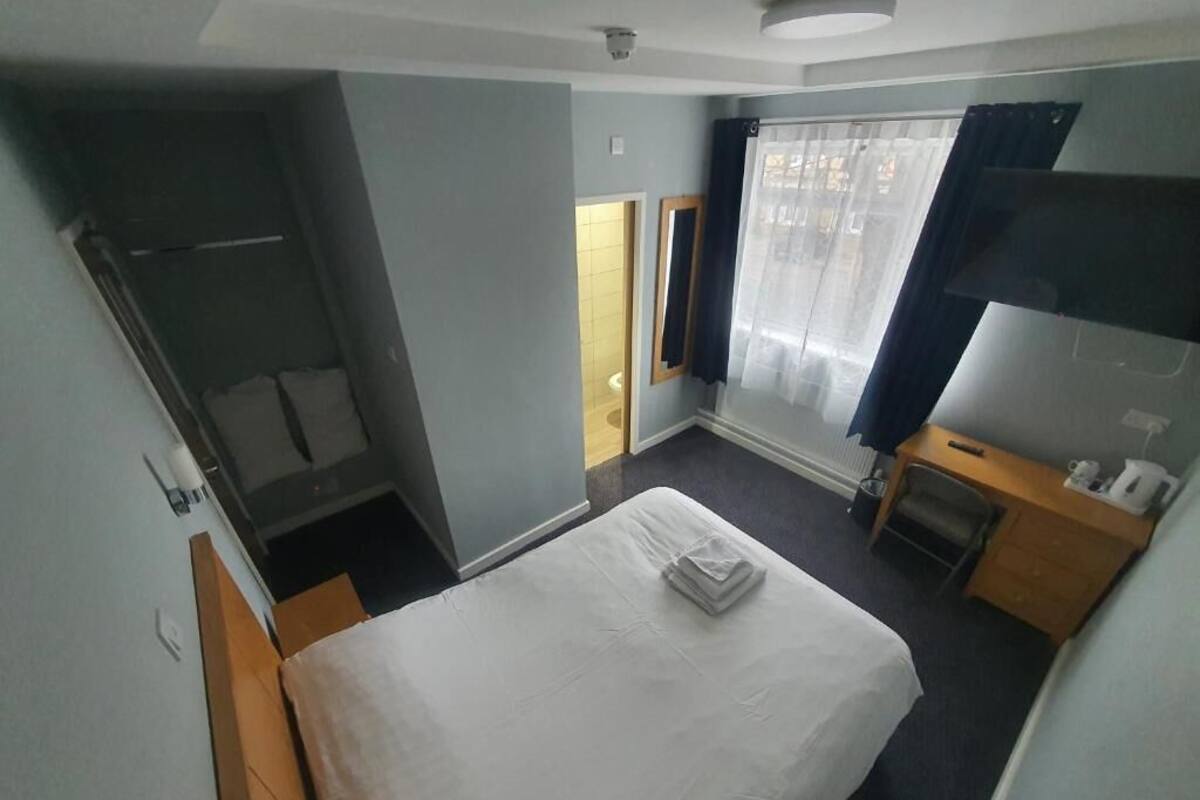 A hotel room with a bed, a small desk with a chair, a mirror, a window with curtains, and a door leading to a bathroom.