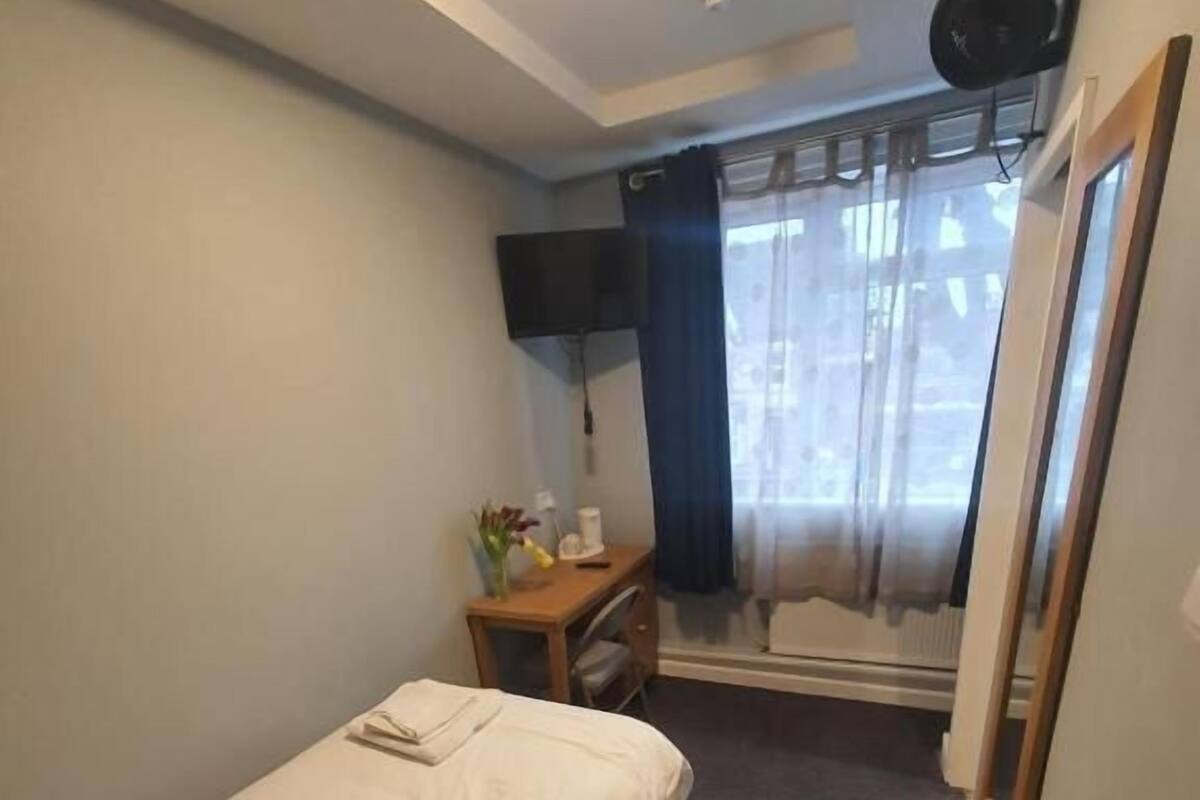 A small, simple room with a single bed, a desk with a chair, a flat-screen TV mounted on the wall, and a window with curtains.