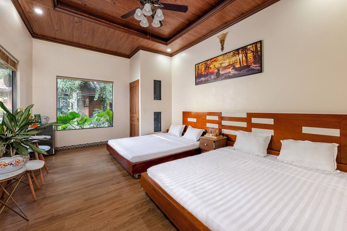 A hotel room with two beds, a ceiling fan, a large window with a view of greenery, and a painting on the wall.