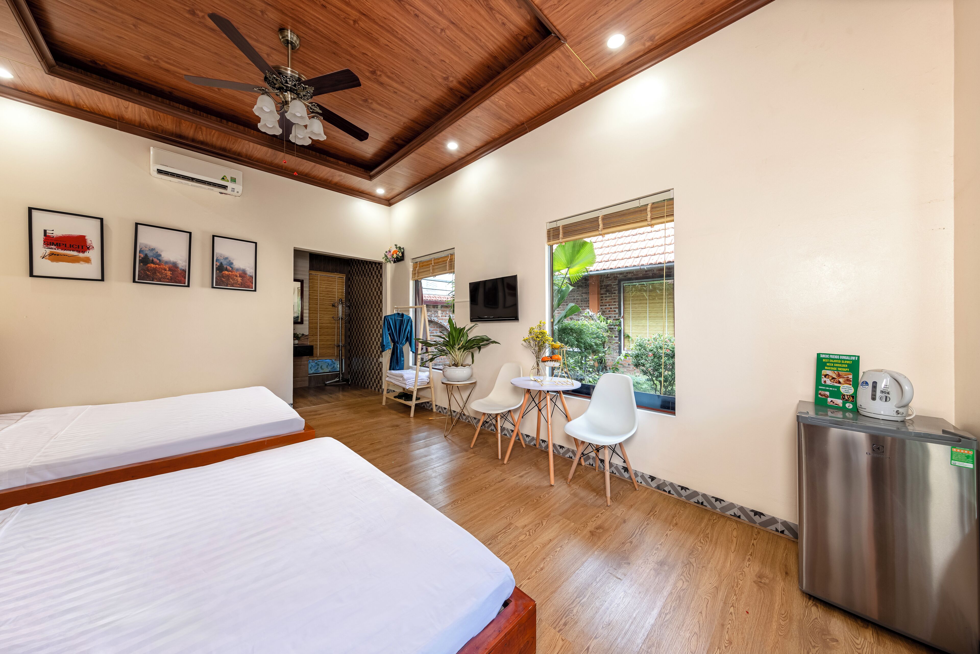 A hotel room with two beds, a ceiling fan, a small table with chairs, a refrigerator, and a window with blinds.