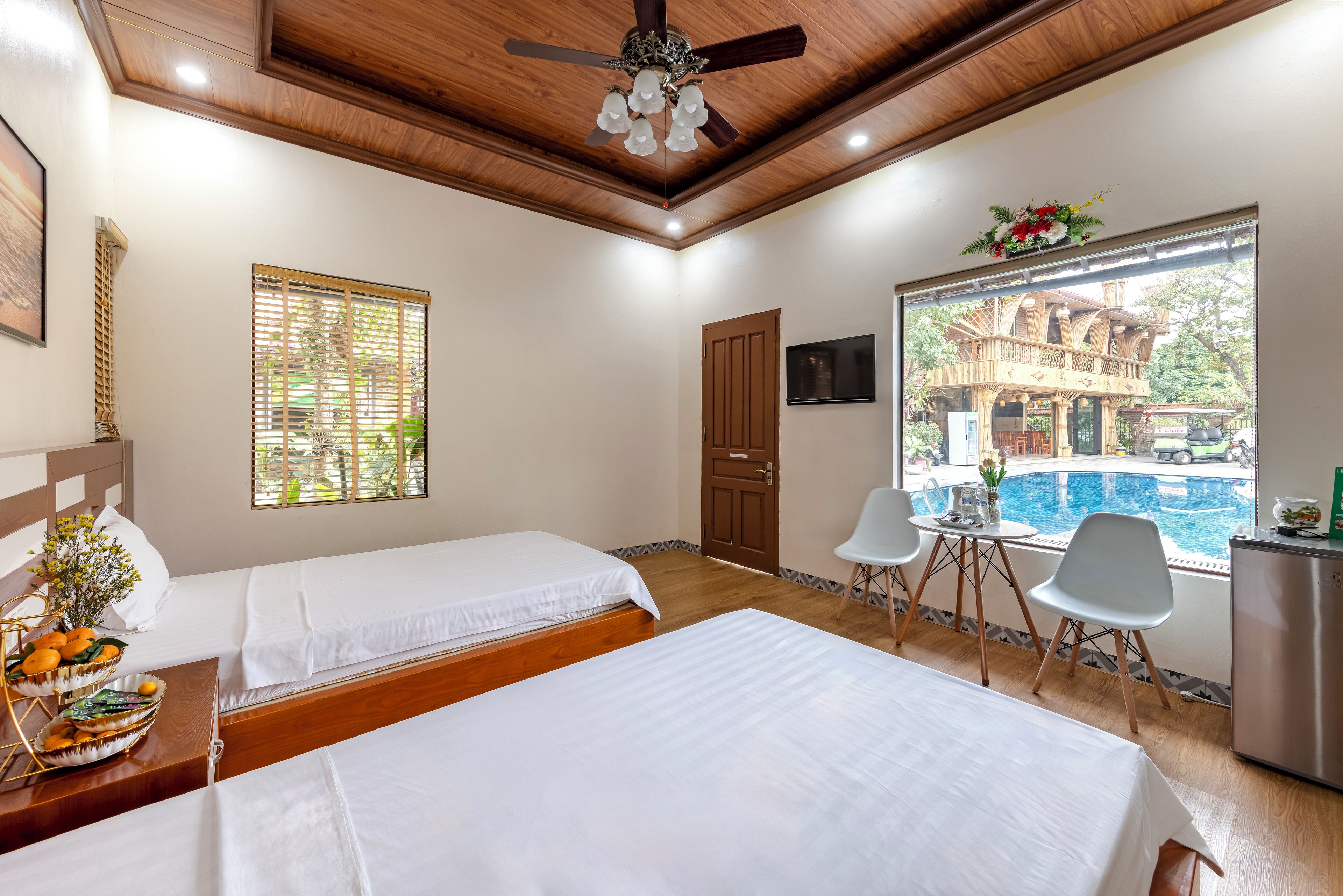 Habitación de hotel con dos camas, ventilador de techo, ventana con persianas, televisión, una mesita con una silla y vista a una piscina al aire libre.