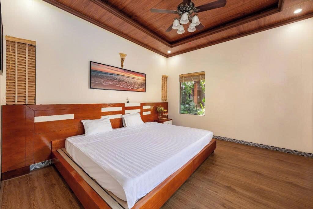 A bedroom with a wooden bed, a ceiling fan, a painting, and a window with a view of greenery.