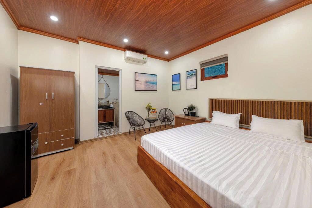 A bedroom with a wooden ceiling, a bed with white bedding, a wardrobe, a desk, two chairs, and a small table with a plant.