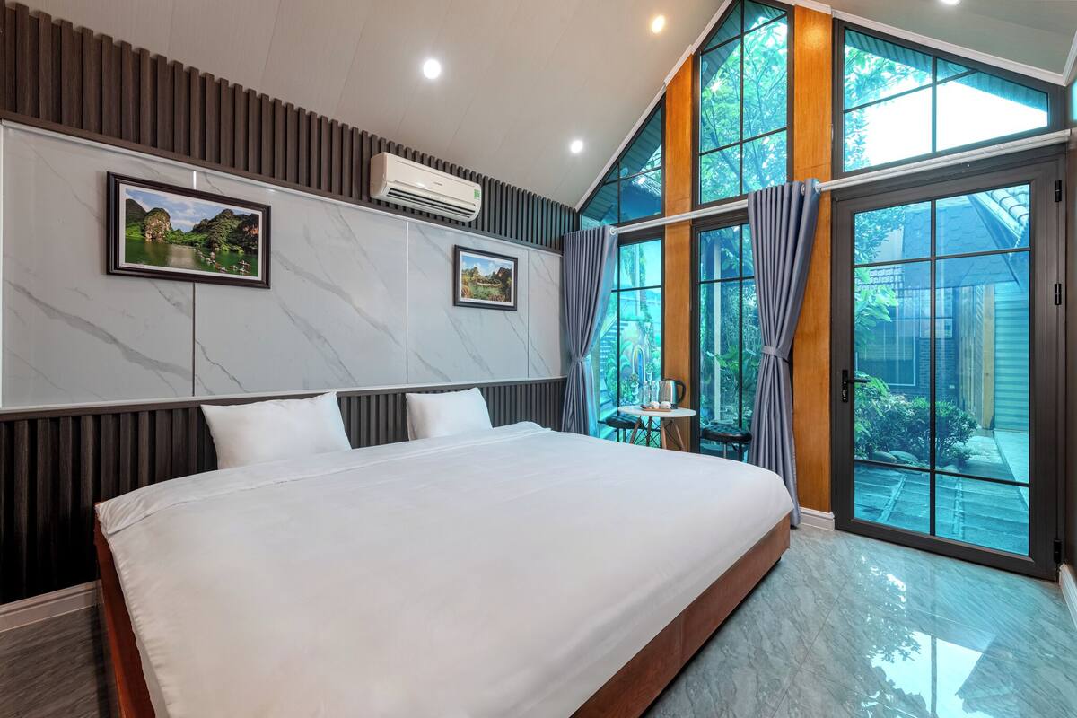 A modern hotel room with a large bed, two framed pictures on the wall, an air conditioning unit, and a view through large glass doors.