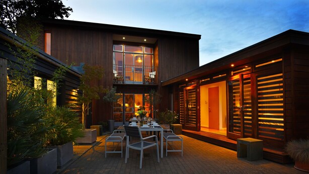 A modern two-story house with a dining area featuring a table and chairs, surrounded by potted plants and a brick patio. - Cedar House at The Old Fire Station (Canterbury)