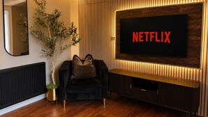 A modern living room with a large flat-screen TV mounted on a wooden wall, a dark-colored sofa, a potted plant, and a radiator. - Cedar House at The Old Fire Station (Canterbury)