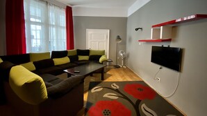A modern living room with a sectional sofa, a flat-screen TV mounted on the wall, a standing fan, and a red and black floral rug. - Casa Balcescu (Brasov)