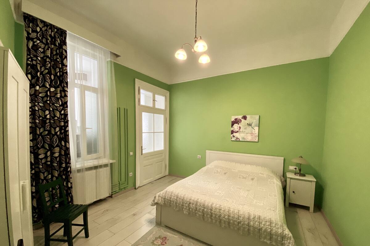 A bedroom with a bed, a nightstand, a lamp, a chair, a window with curtains, and a wall with a framed picture.