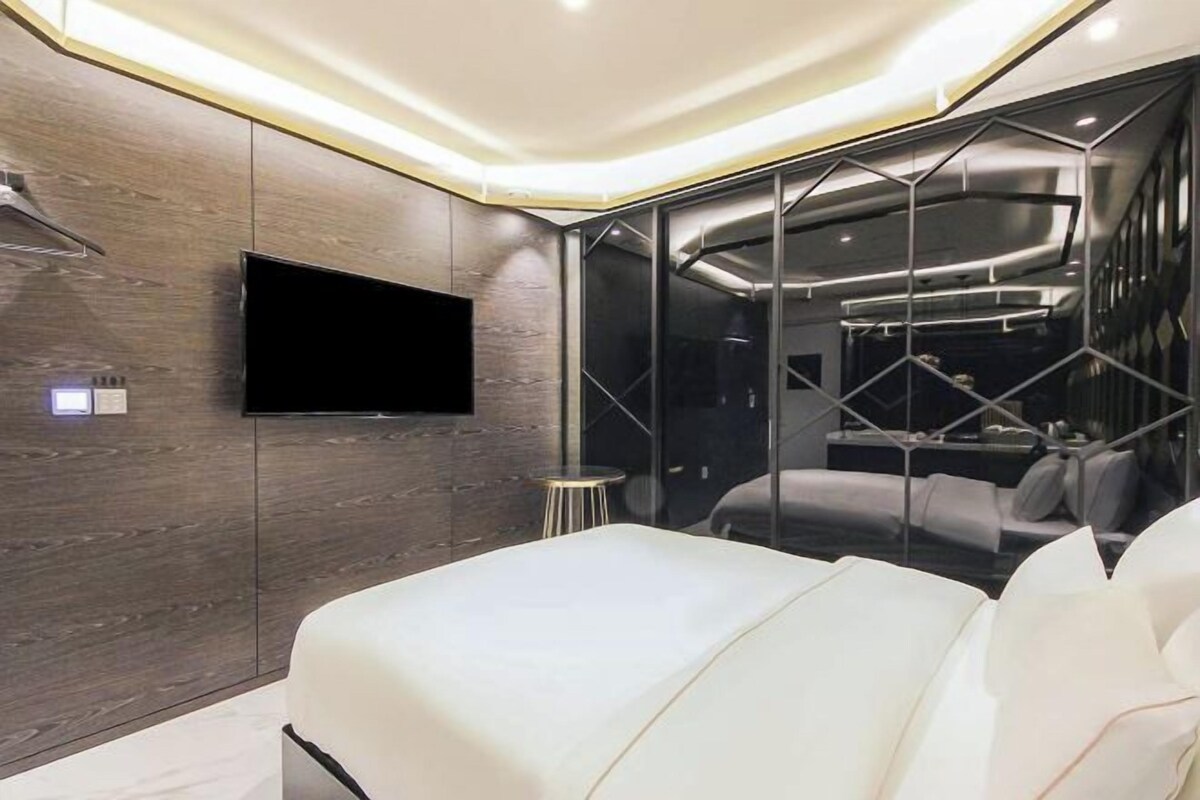Un dormitorio moderno con una cama grande, un televisor de pantalla plana montado en la pared y un panel de vidrio que separa la habitación.