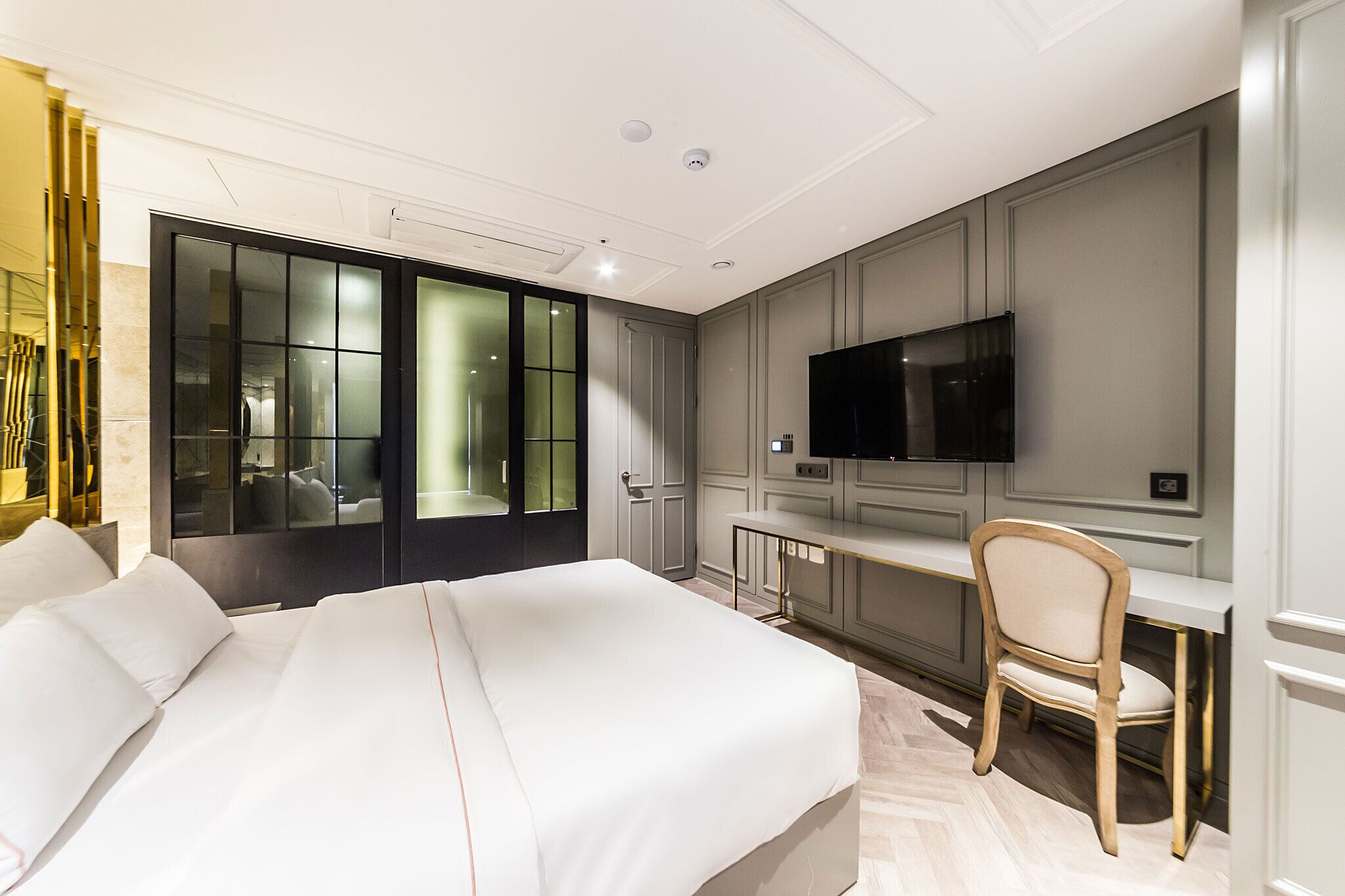A modern hotel room with a large bed, a desk with a chair, a television, and a sliding door to the bathroom.