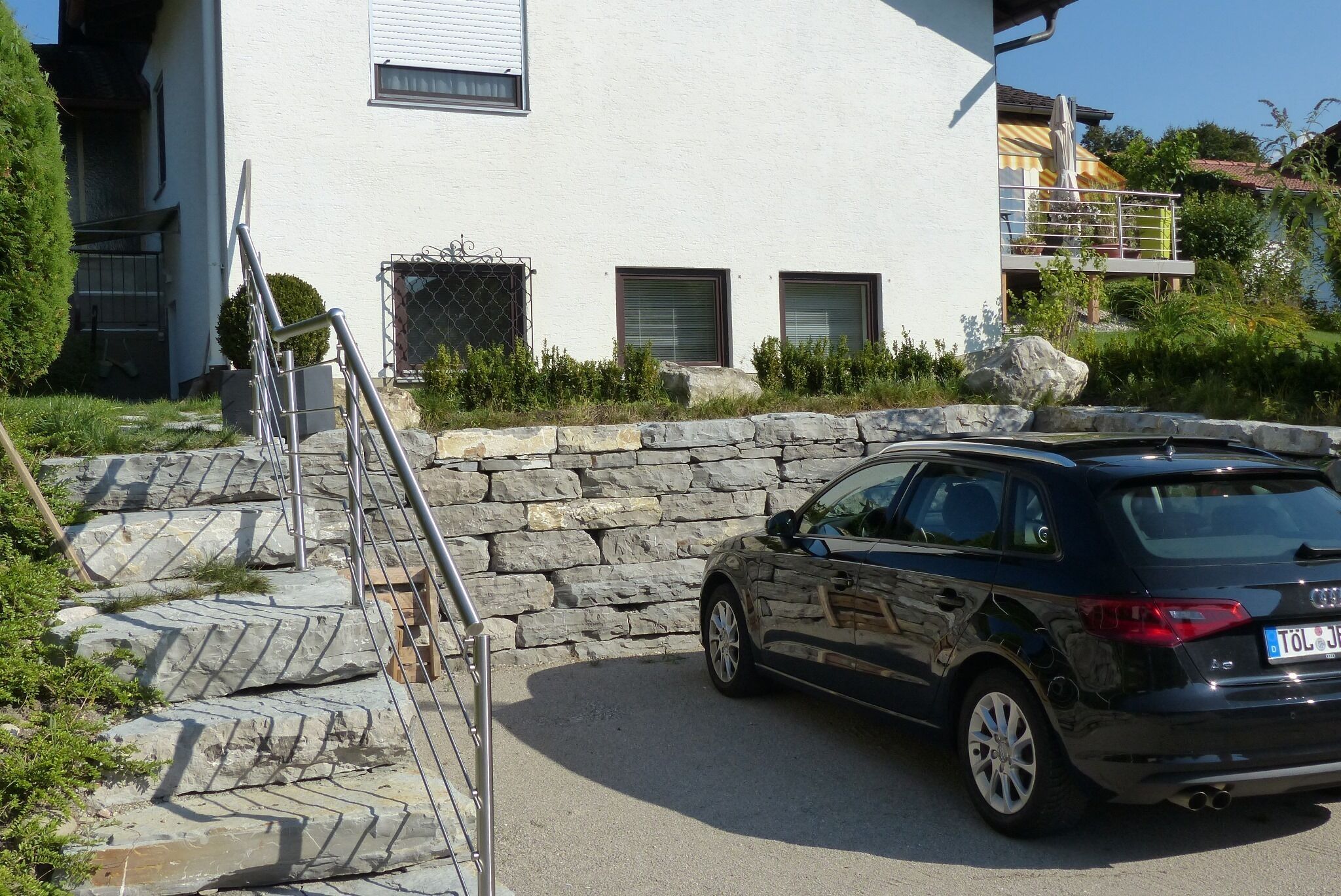 Une voiture noire est garée devant un bâtiment blanc, doté d’un mur en pierre et d’une rambarde en métal.