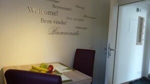 Una habitación con una pared que muestra letreros de bienvenida en varios idiomas, una mesa de comedor con una cesta de frutas y una puerta con un cartel encima.