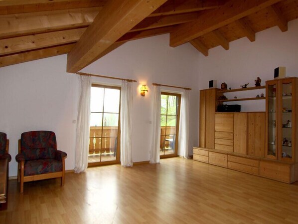 A spacious living room with wooden beams, a large window with curtains, a wooden cabinet with glass doors, and a patterned armchair.