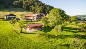 Un chalet grande de varios pisos con techo rojo, rodeado de campos verdes exuberantes y una colina boscosa al fondo.