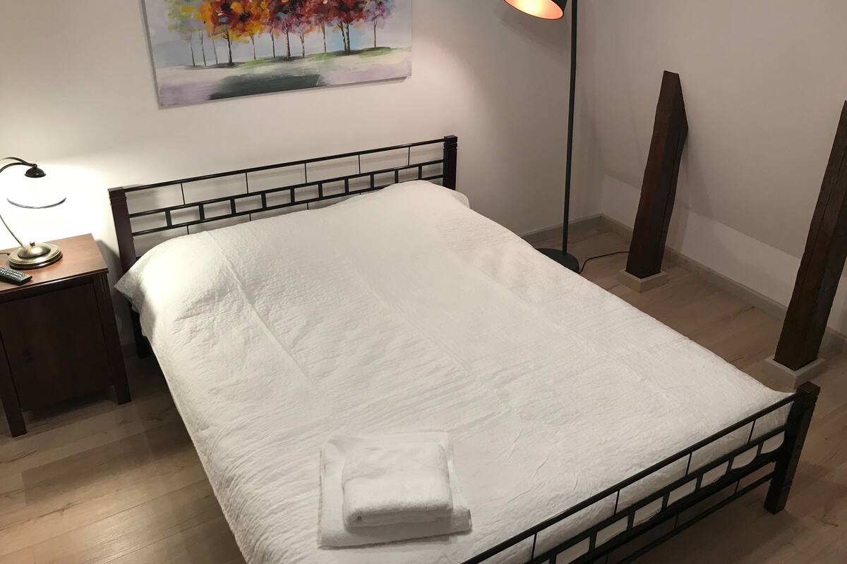 Ein ordentlich bezogenes Bett mit weißer Decke und einem gefalteten Handtuch, eine Nachttischlampe, ein Baumgemälde und eine Stehlampe.