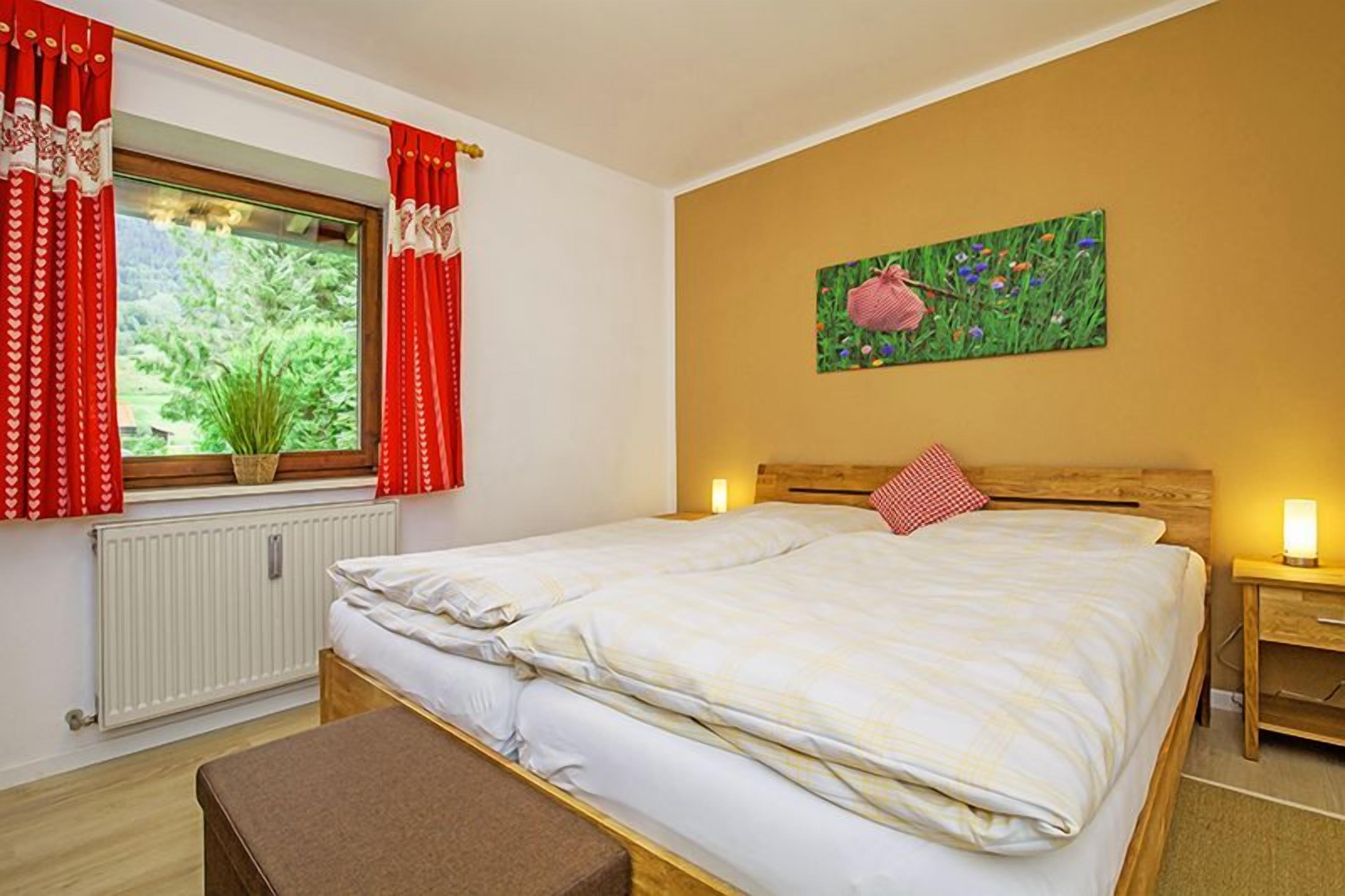 A bedroom with a double bed, a window with curtains, a radiator, a bedside table with a lamp, and a painting on the wall.