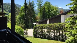 Vue depuis l’intérieur d’un bâtiment, donnant sur un jardin bien entretenu, avec une variété d’arbres et une montagne en arrière-plan.