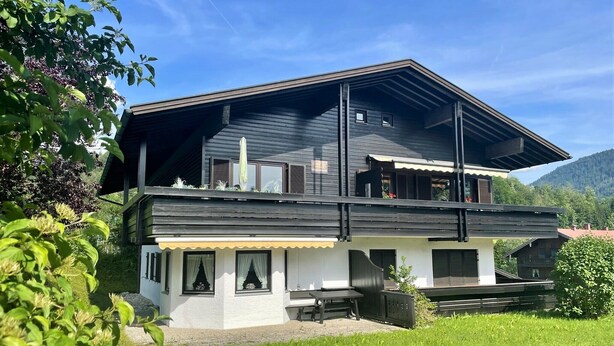 Une maison à deux étages avec un balcon, un jardin et un paysage montagneux en arrière-plan.