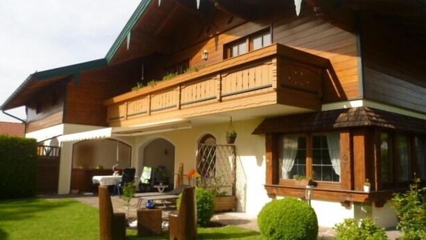Uma casa de madeira de dois andares, com varanda, um pequeno alpendre e um jardim bem cuidado.
