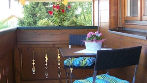 Um deck de madeira com mesa e cadeiras, um lustre pendurado e uma janela com vista para o verde.