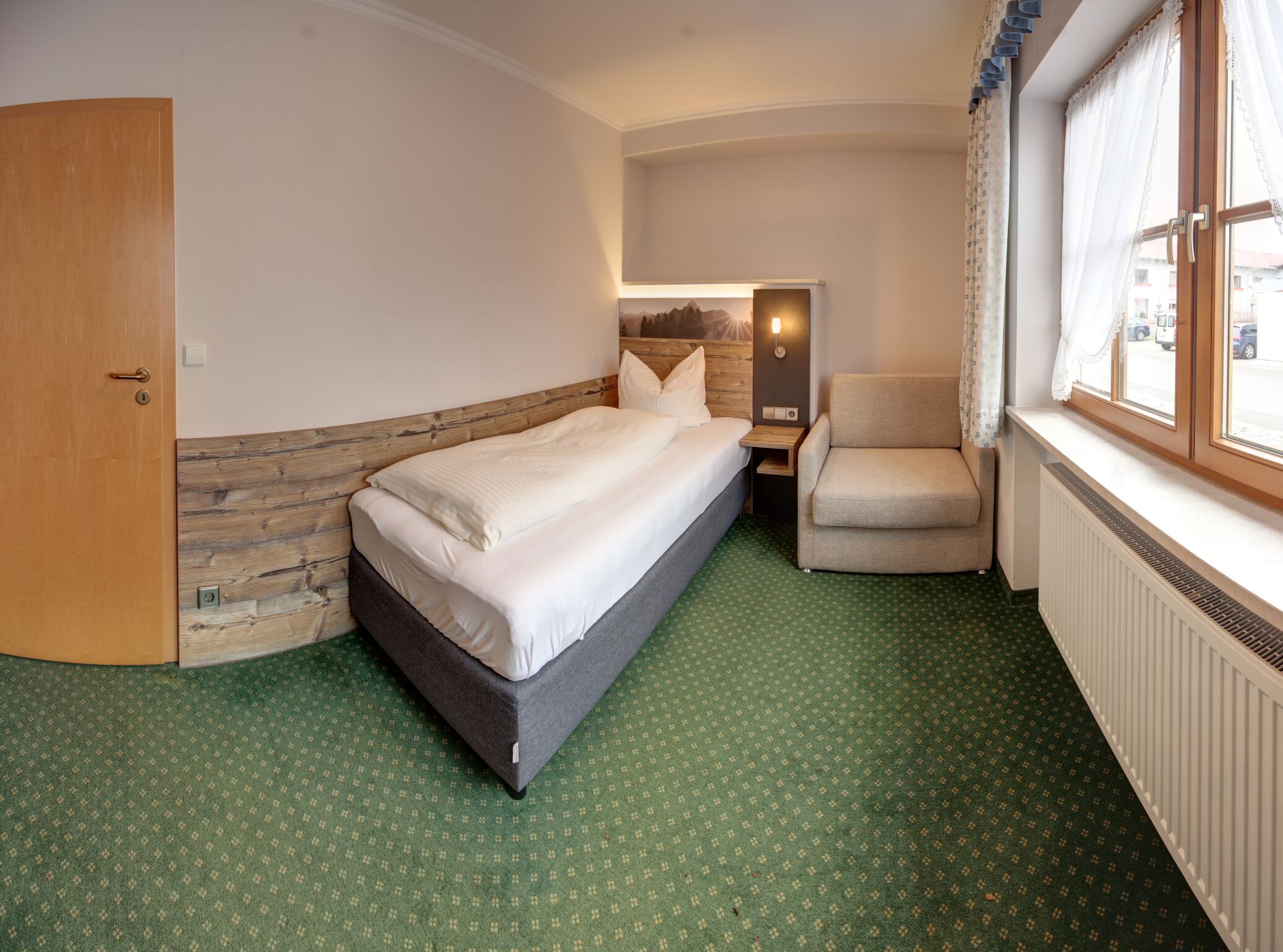 A hotel room with a bed, a chair, a window with curtains, a radiator, and a door.
