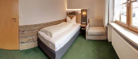 A hotel room with a bed, a chair, a window with curtains, a radiator, and a door.