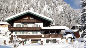 A chalet-style building with a wooden facade and green shutters, surrounded by snow-covered trees and a clear sky.