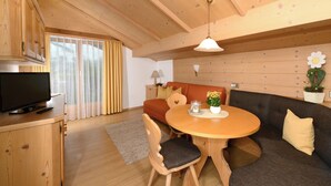 A cozy room with a wooden ceiling, a television on a cabinet, a round dining table with chairs, a sofa, and a small potted plant.