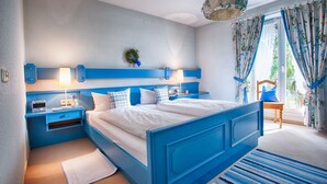 A bedroom with a blue bed, bedside tables with lamps, a small rug, and a view of the outdoors through a window with curtains. - Stylish apartment with beautiful mountain scenery (St5)/Steilhofweg. 65 sqm, south-facing balcony (Bayerisch Gmain)