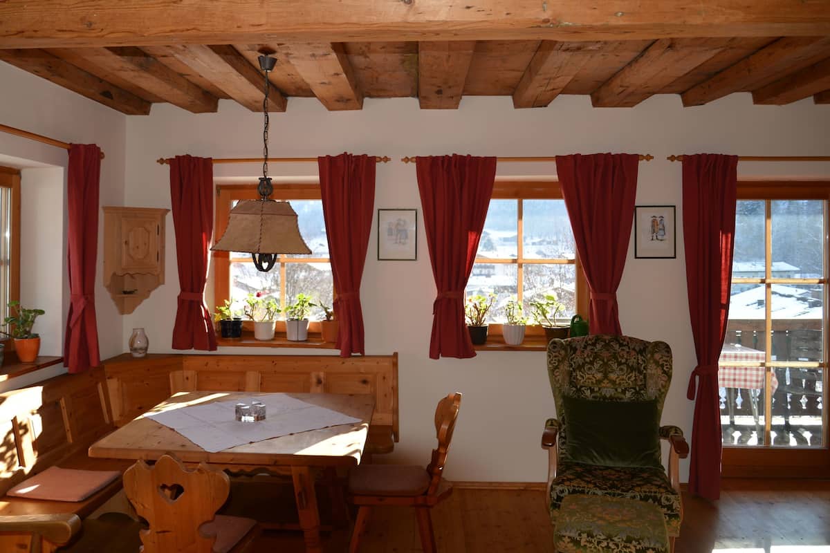 Ein gemütliches Zimmer mit Holzbalken, einem Esstisch mit Stühlen, einem gepolsterten Sessel und Fenstern mit roten Vorhängen.